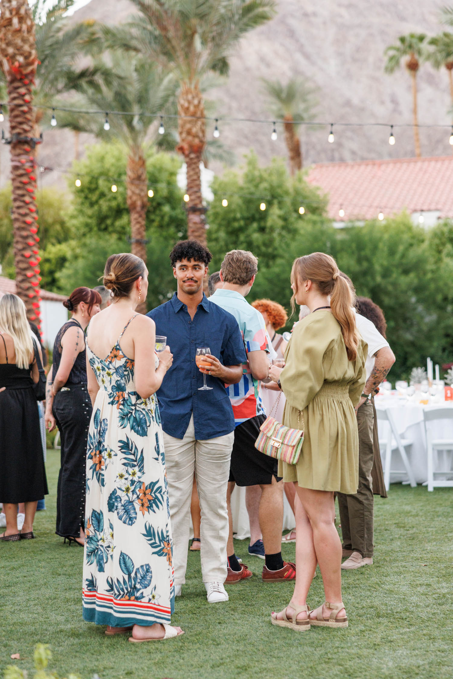 People stand and talk in small groups at an outdoor event on grass, holding drinks. Palm trees, string lights, and mountains are visible in the background.