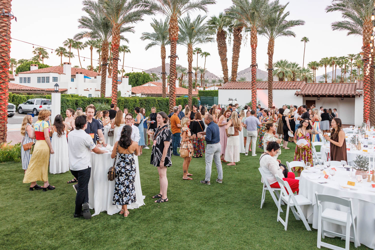 A large group of people socialize outdoors among palm trees, standing and sitting at round tables set with white tablecloths on a grassy lawn.