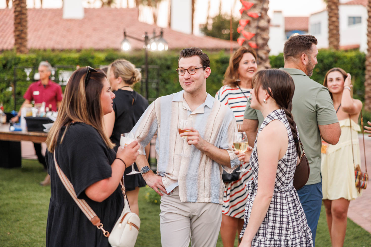 People stand outdoors in a group, holding drinks and conversing, with palm trees and buildings in the background.