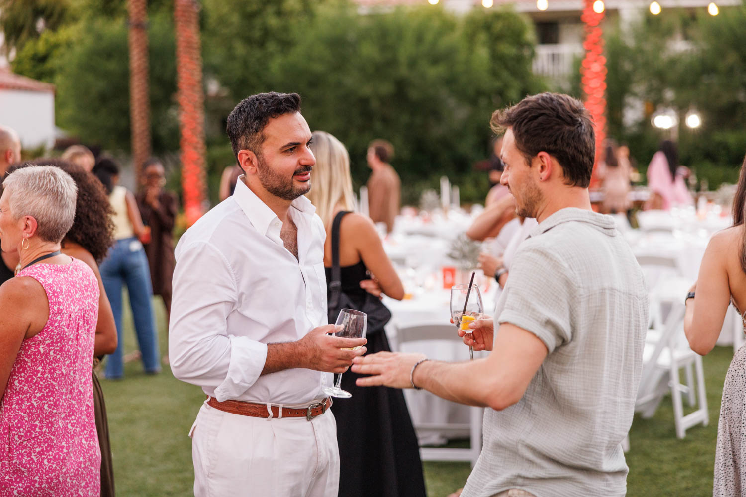 Two men stand outdoors talking, each holding a drink, surrounded by other people at an event with round tables and white chairs.