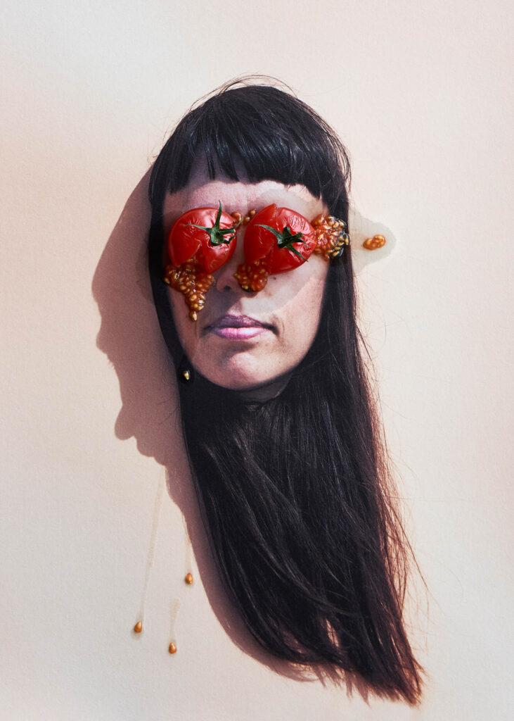 A woman’s head with long dark hair lies on a pale surface, with two tomatoes and seeds covering her eyes.