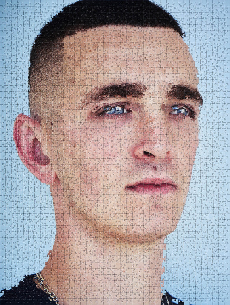 A completed jigsaw puzzle forms the image of a young man with short hair, blue eyes, and a chain necklace, set against a light blue background.