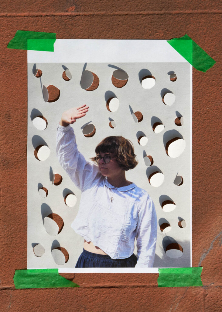 A photo of a person in glasses and a white shirt standing behind a perforated paper sheet taped to a textured brown wall. The person raises one arm.