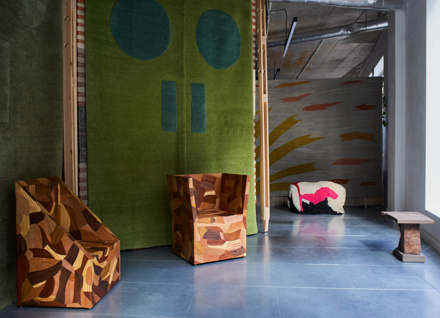 Modern room with geometric wood armchairs, green wall tapestry, abstract rug, low table, and concrete ceiling, illuminated by natural light.