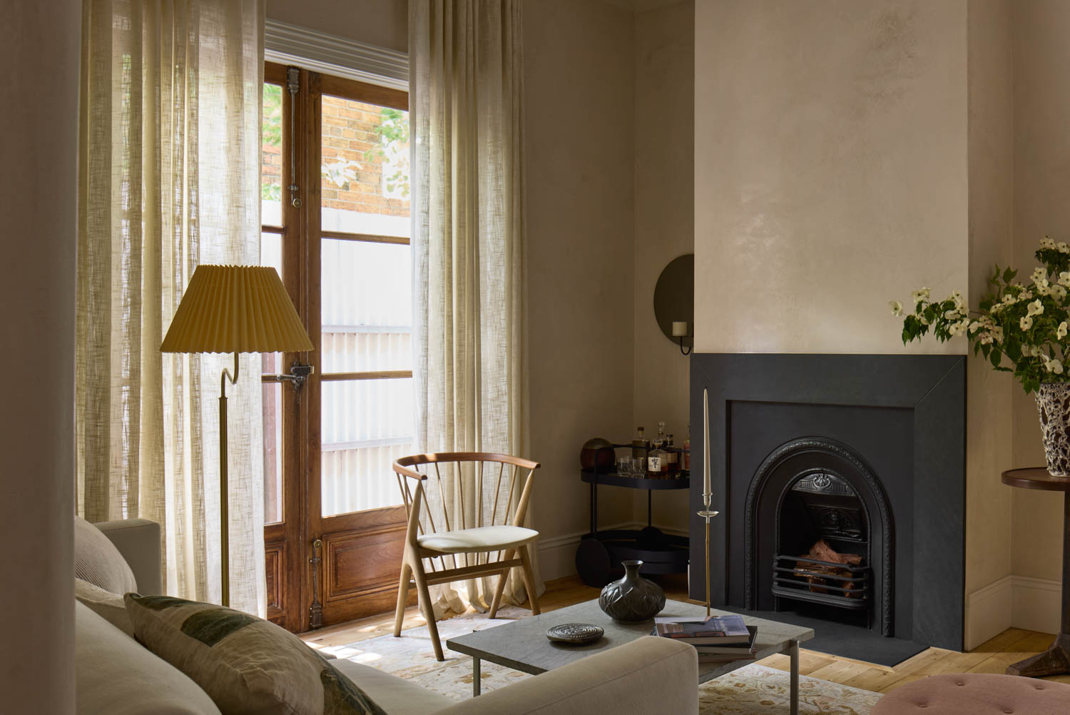 A living room with a fireplace, a wooden chair, a sofa, a standing lamp, a round side table with flowers, and sheer curtains covering glass doors.