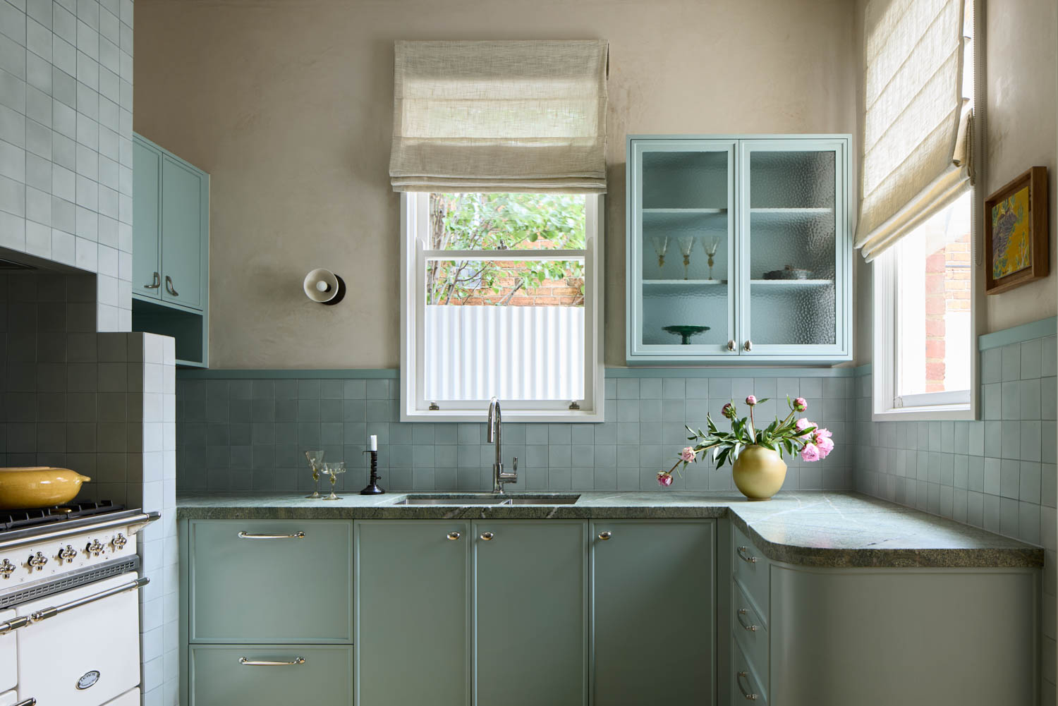 Light blue kitchen with tiled backsplash, glass-front cabinets, and a vase of flowers on the counter. Two windows with beige Roman shades let in natural light.