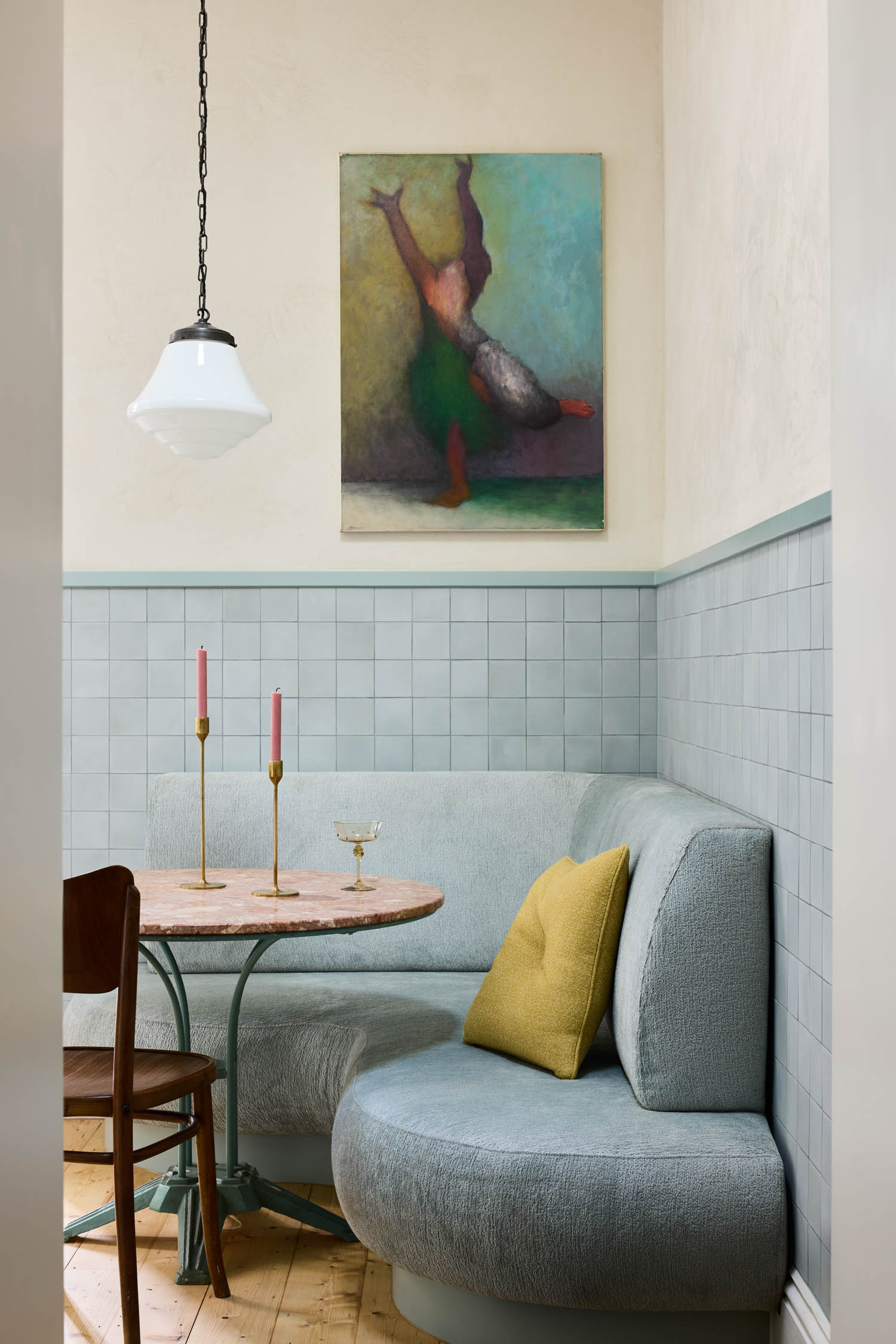 A cozy dining nook with a curved blue-gray banquette, round table, wood chair, two pink candles, a yellow pillow, and a painting of a dancer on the wall above.
