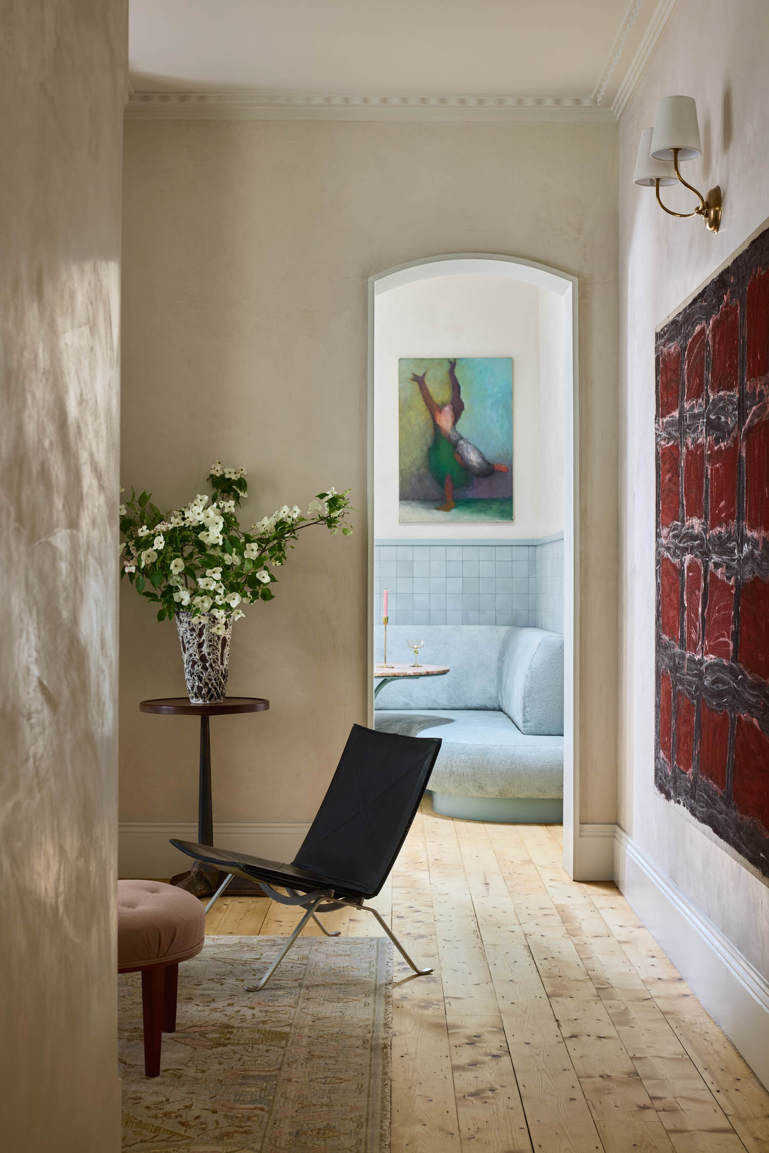A hallway with light wood floors, a black chair, floral arrangement, large abstract painting, and an arched doorway leading to a pastel-colored room with a sofa and artwork.