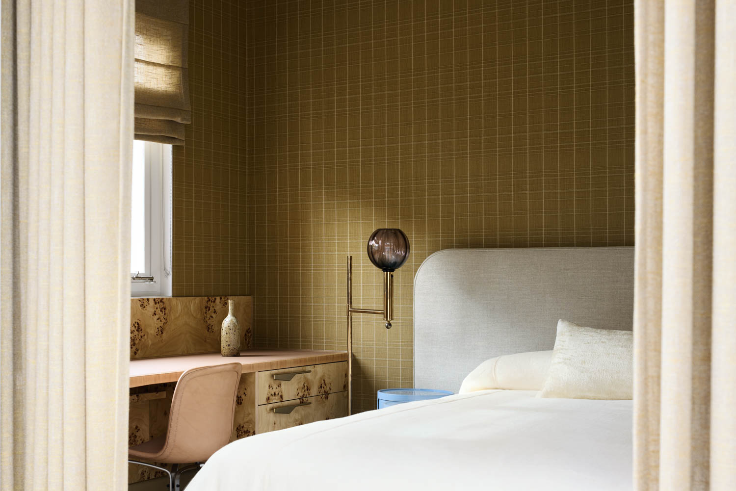 A modern bedroom with a light upholstered bed, a small blue side table, a desk with a pink chair, and yellow grid-patterned wallpaper.
