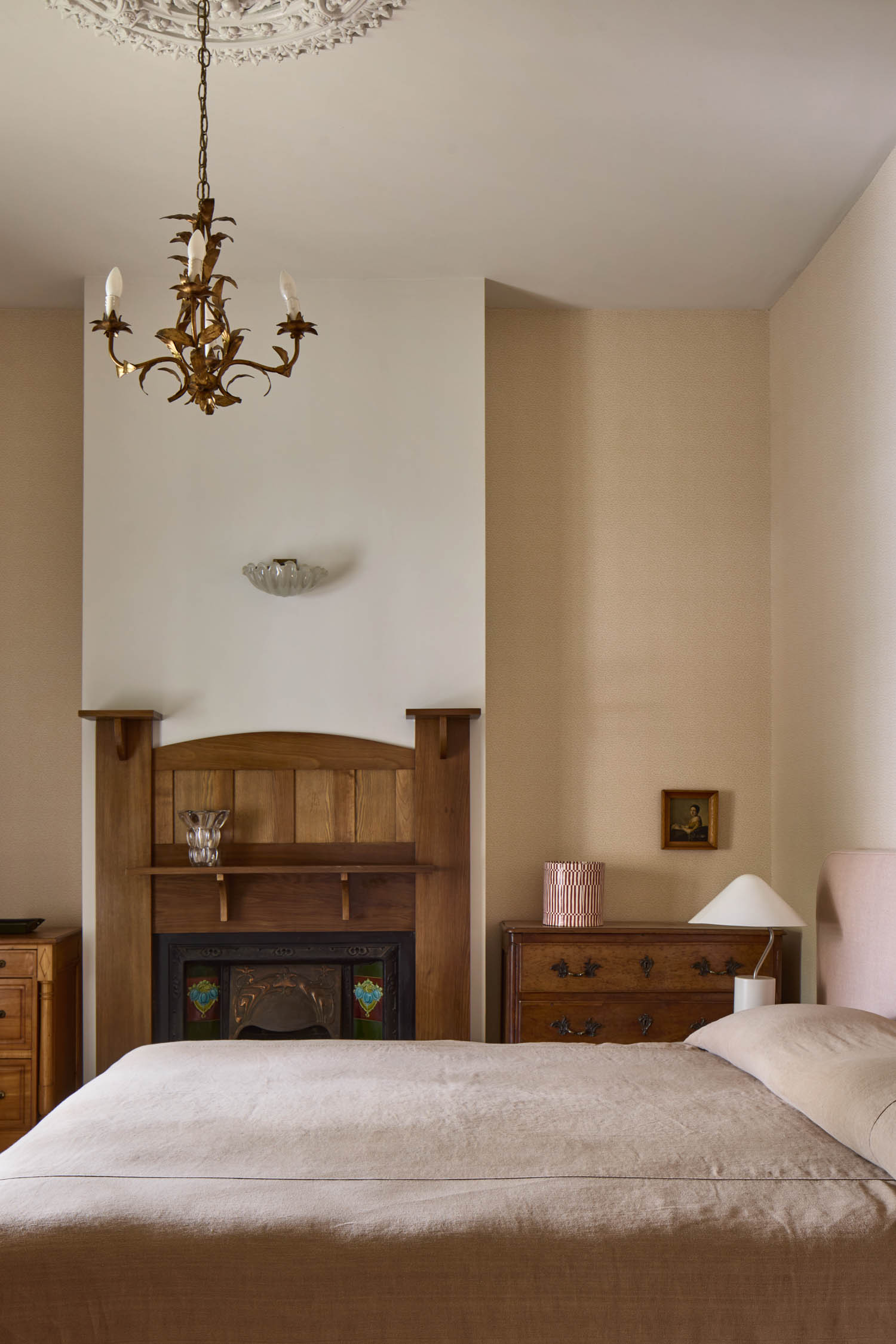 Bedroom with a beige bed, wooden dressers, ornate fireplace, chandelier, table lamp, and light-colored walls.