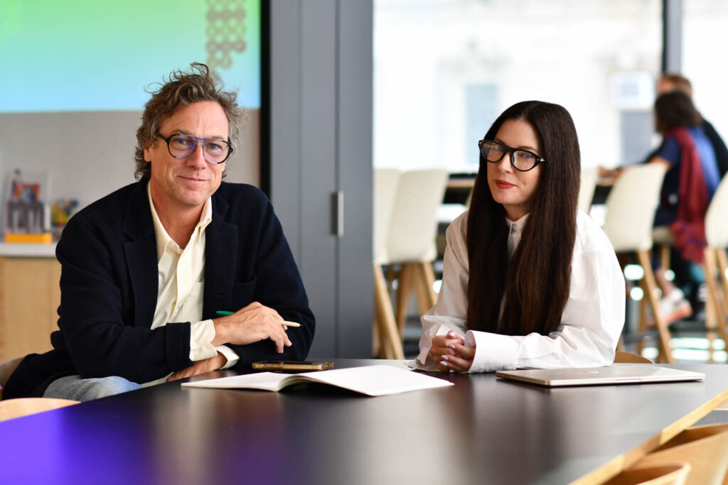 Two people sit at a table with notebooks and a laptop, facing the camera in a modern office setting.