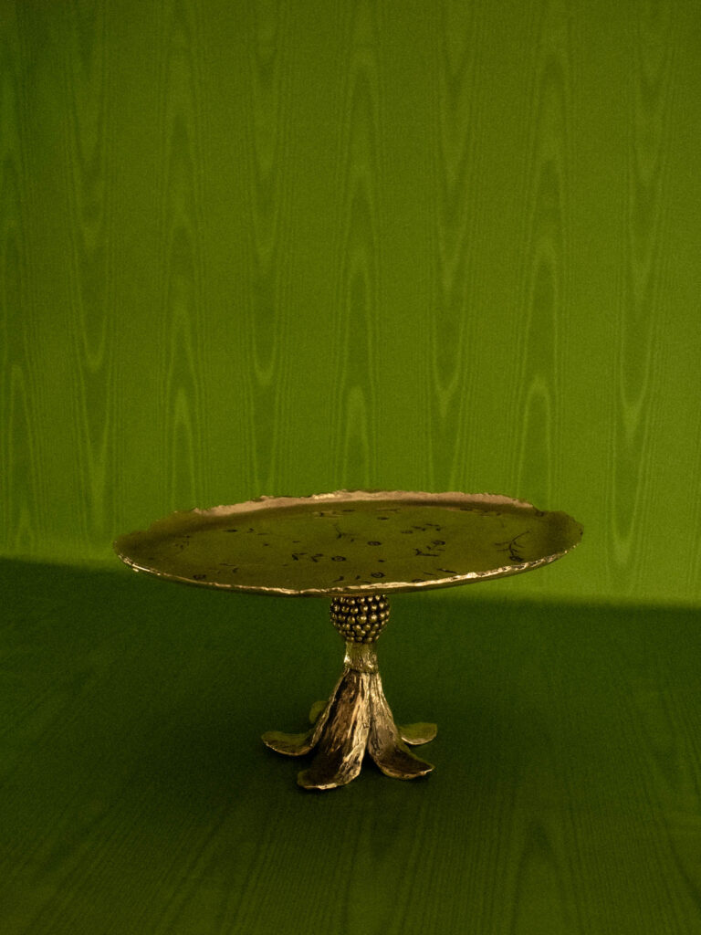 A metallic cake stand with a textured, uneven edge and a sculpted floral base sits on a green surface against a green patterned background.
