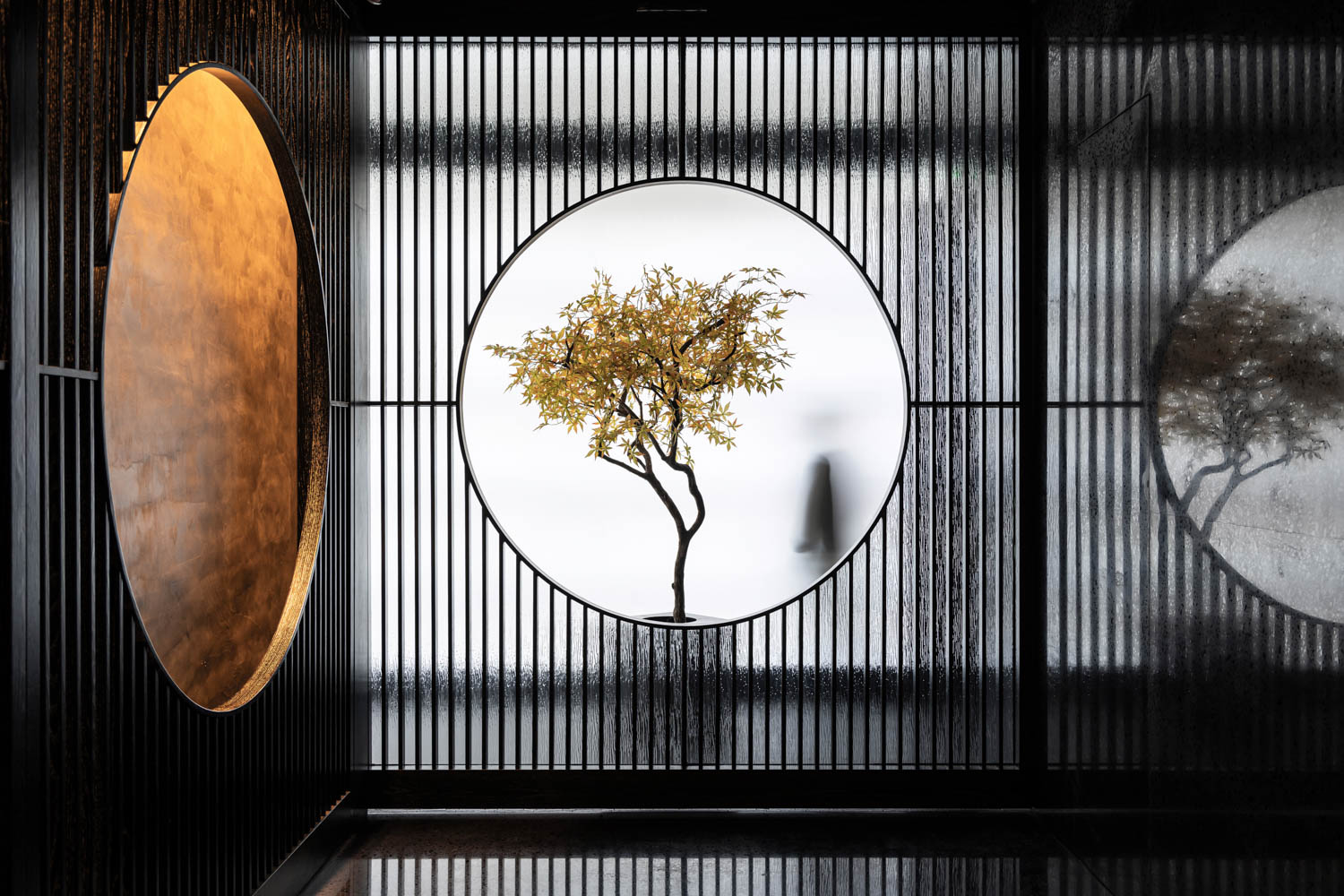 A small tree is framed by a circular window with vertical slats; a blurred figure walks in the background, creating a minimalist, modern scene.