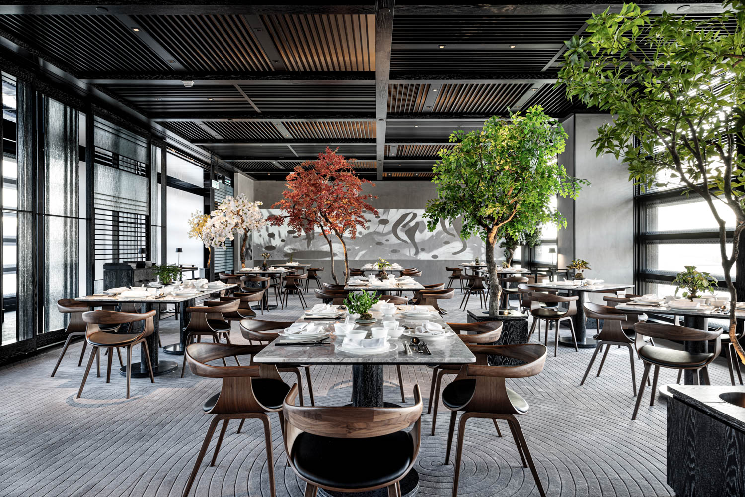 Modern restaurant dining area with wooden tables and chairs, decorated with large indoor trees and natural light from tall windows.