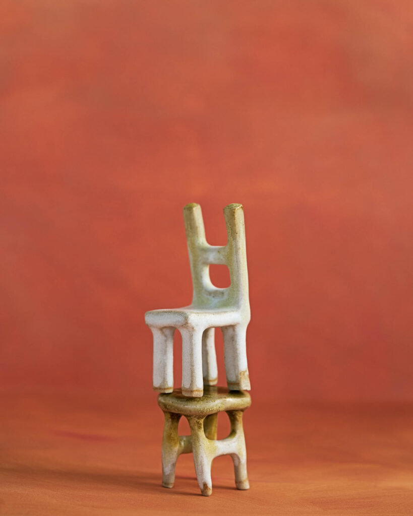 A small ceramic chair is stacked on top of a matching ceramic stool, both with abstract shapes, against an orange background.