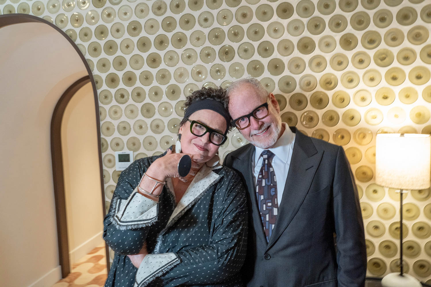 Two people wearing glasses stand close together in a room with patterned wallpaper, one in a dark robe holding glasses, the other in a suit, both smiling at the camera.