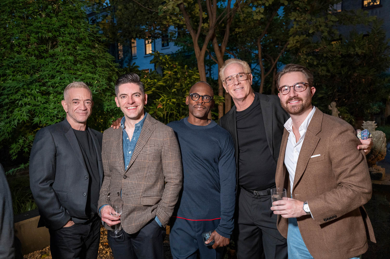 Five men stand together outdoors at an evening event, smiling at the camera. Some hold drinks. Trees and greenery are visible in the background.