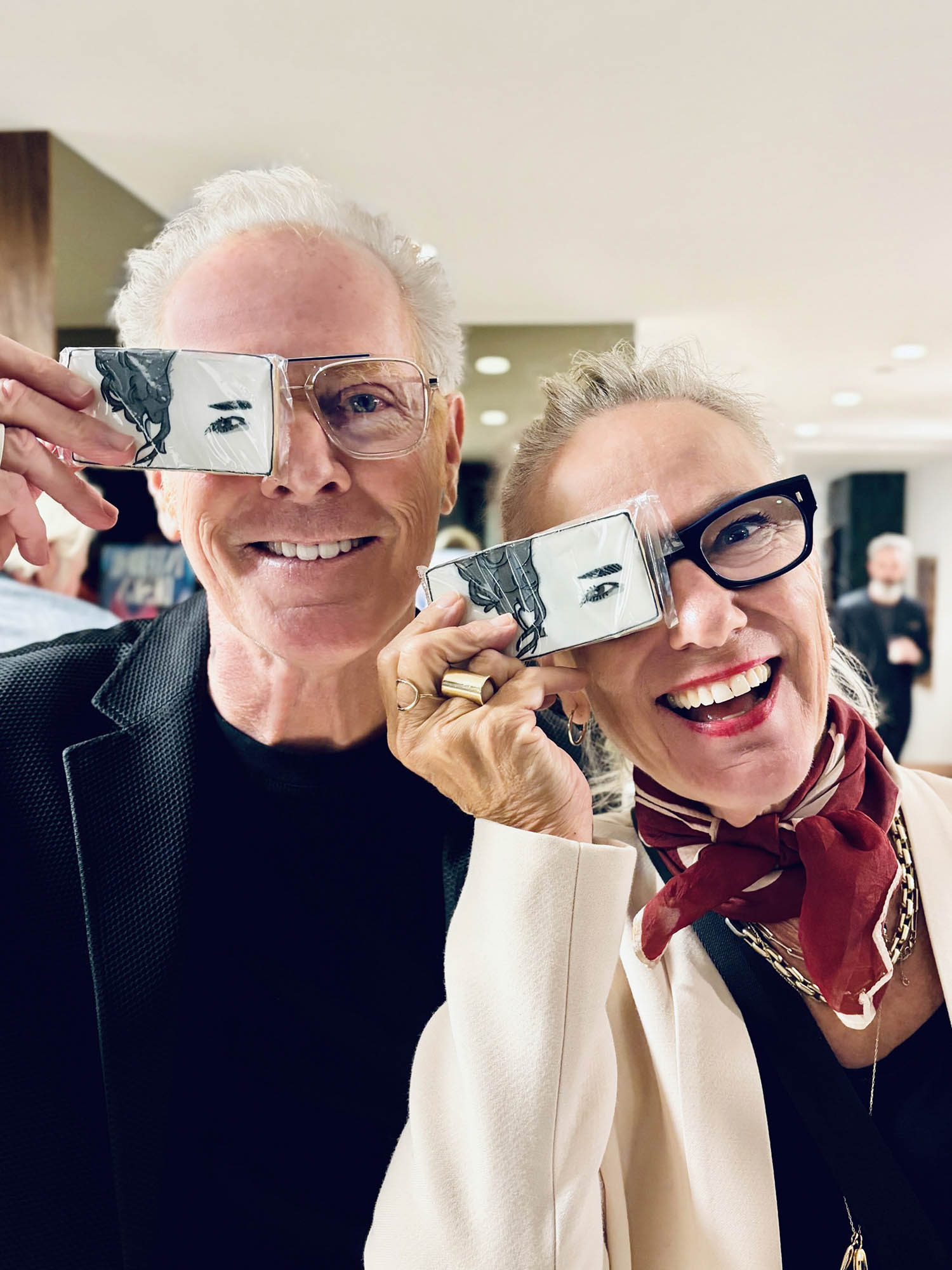 Two smiling adults hold cards with sketched eyes over one of their own eyes, standing indoors and posing playfully for the camera.