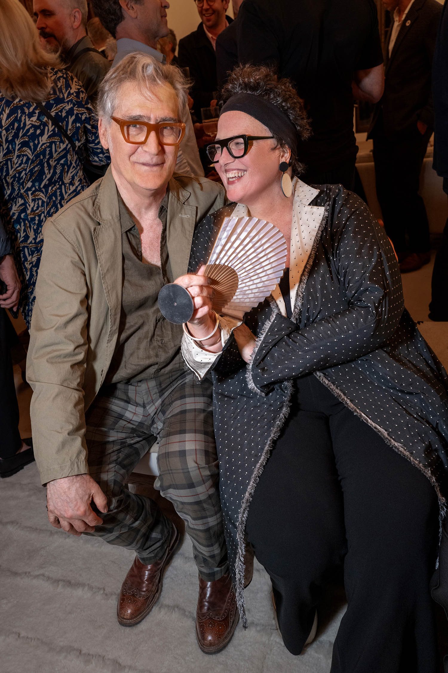 A man in plaid pants and glasses sits beside a woman in black holding a handheld fan, both smiling at an indoor social gathering.