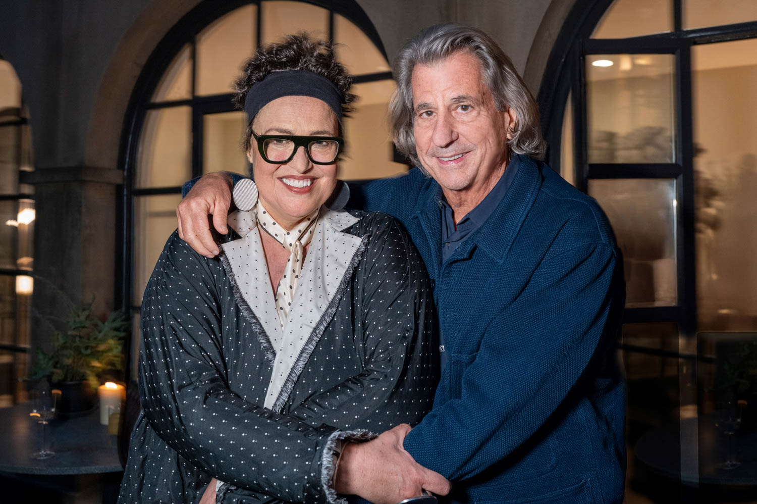 Two adults stand close together, smiling at the camera. One wears glasses and a polka-dot scarf; the other has gray hair and a blue jacket. They are outdoors near a building with large windows.