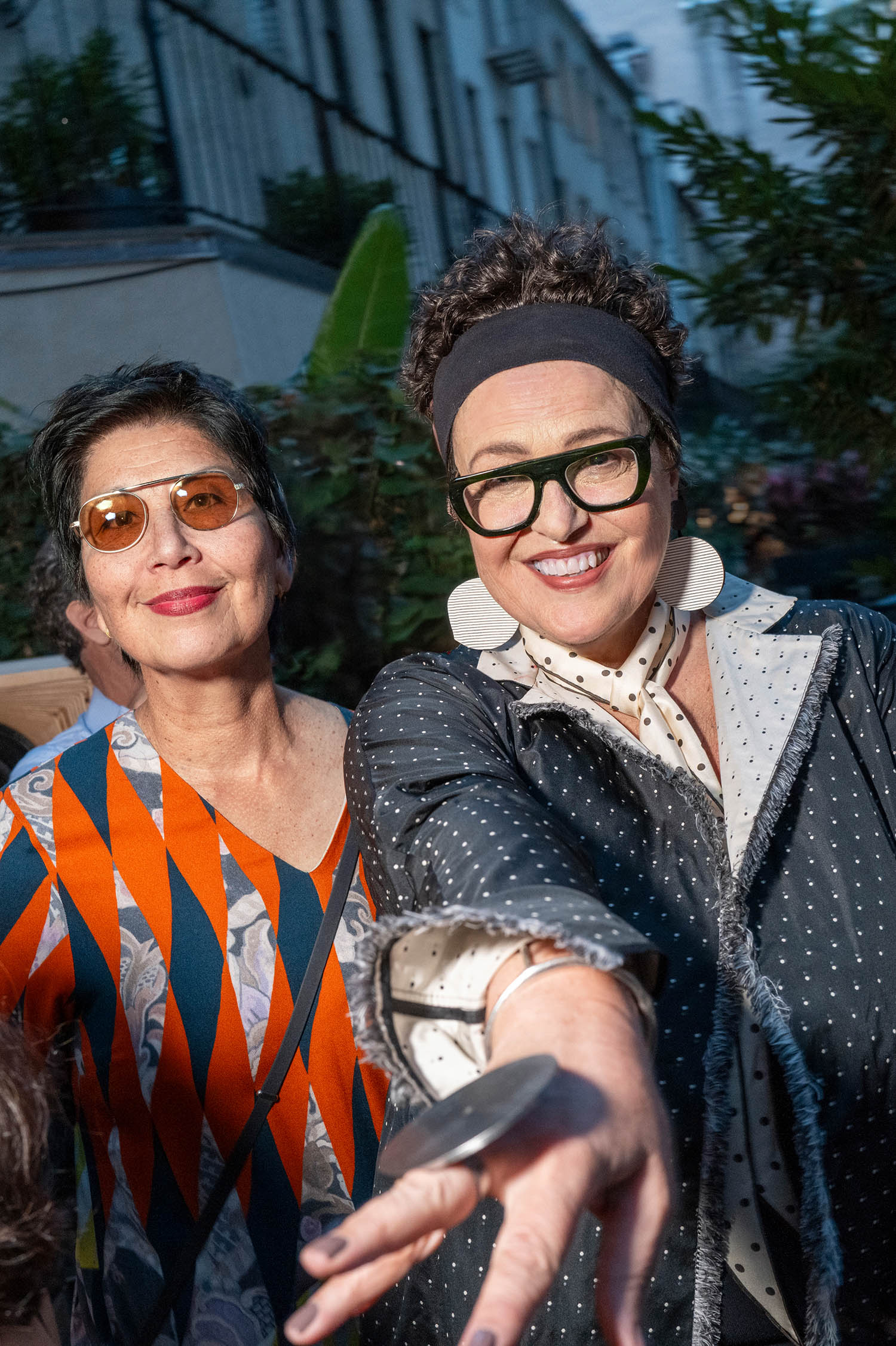 Two women standing outdoors, smiling at the camera. One wears round glasses and patterned dress, the other wears large glasses, earrings, and a polka-dot coat, extending her hand forward.