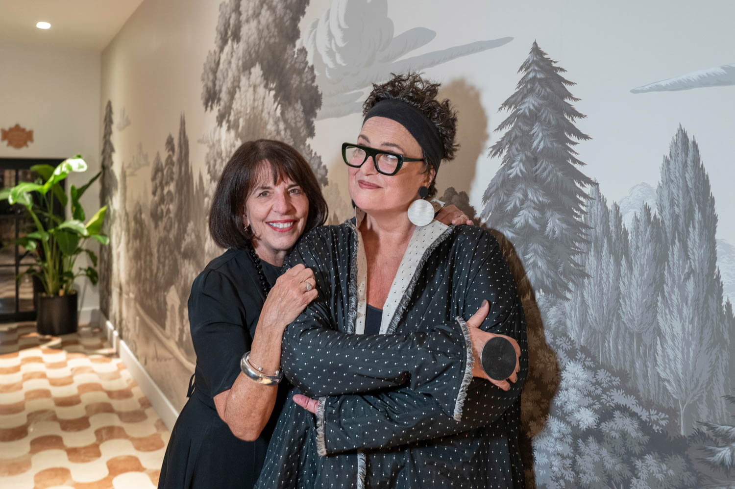 Two women stand together in a hallway with one woman’s arm around the other, smiling. The background features a mural of trees and a potted plant in the corner.