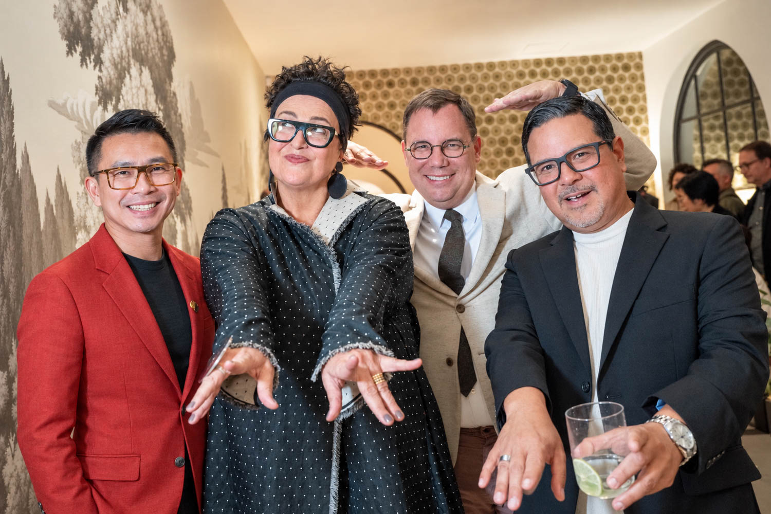Four adults pose indoors at a social event; two gesture playfully with their arms while the others stand smiling, one holding a drink.