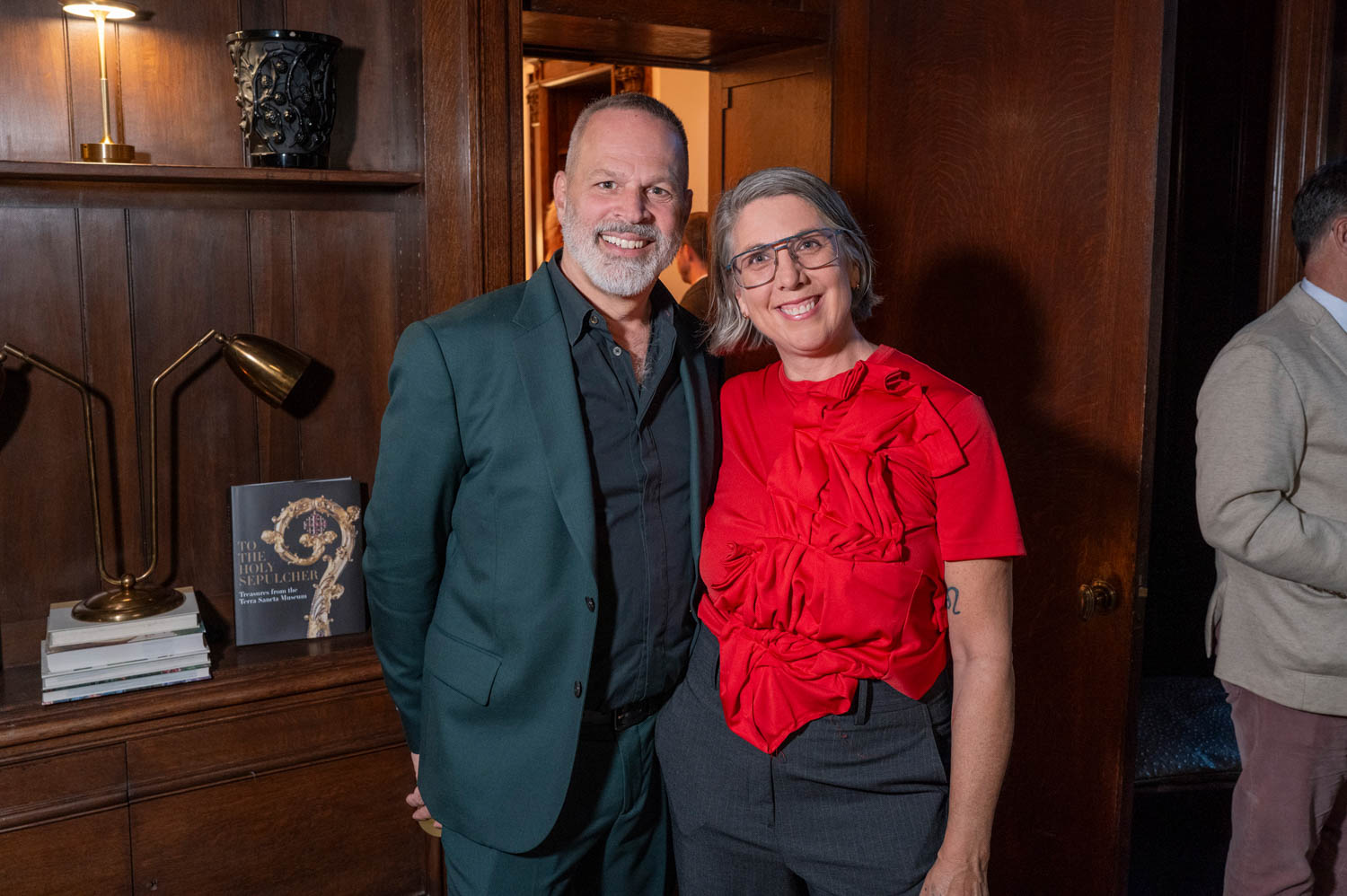 A man in a dark suit and a woman in a red blouse stand side by side, smiling, in a wood-paneled room with books and a lamp on the shelf behind them.