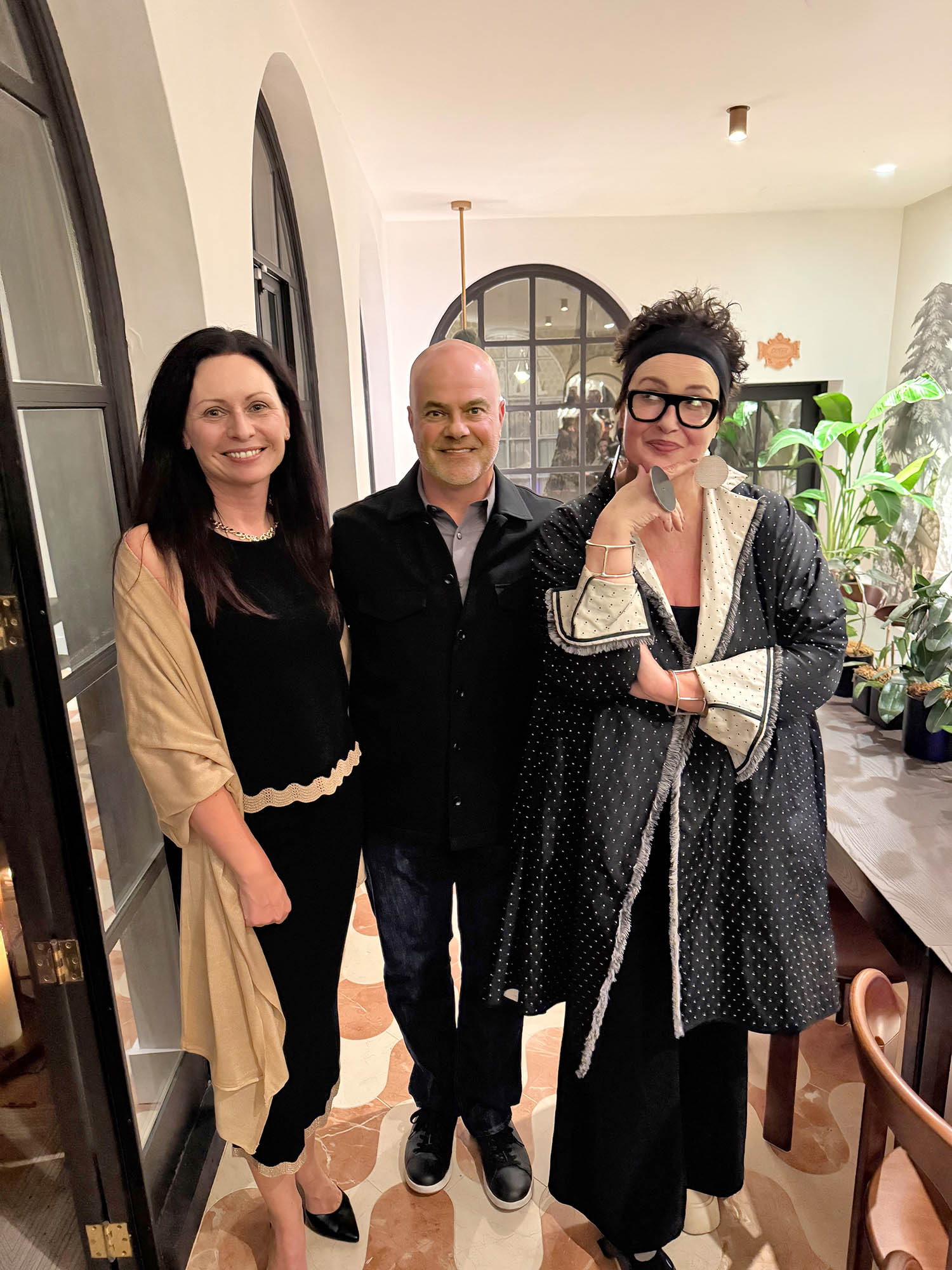 Three adults stand together indoors, smiling at the camera. The room has arched windows, plants, and patterned flooring. One person wears glasses and a polka-dot outfit.
