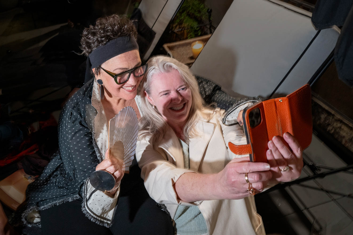 Two women smile and pose for a selfie; one holds an orange phone and the other holds a translucent award. Both appear happy and celebratory.