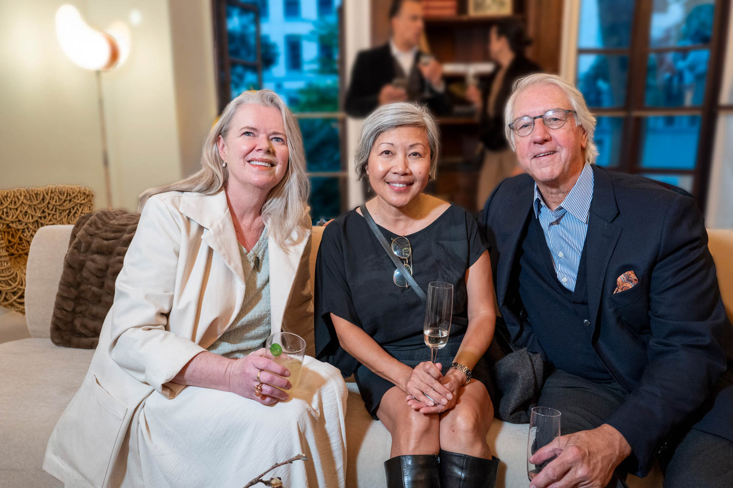 Three adults sit on a couch at a social event, smiling at the camera and holding drinks. Two people are blurred in the background near a window.