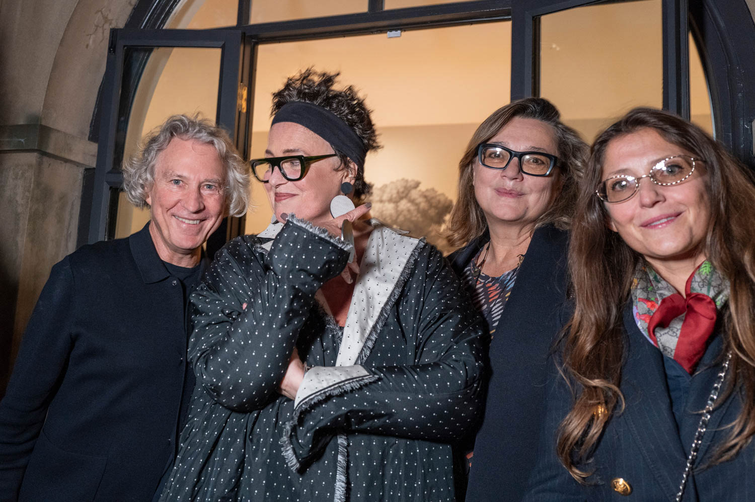Four adults stand closely together, posing for a photo indoors at night. Three wear glasses and one wears large earrings and a polka dot coat. All are looking toward the camera and smiling.