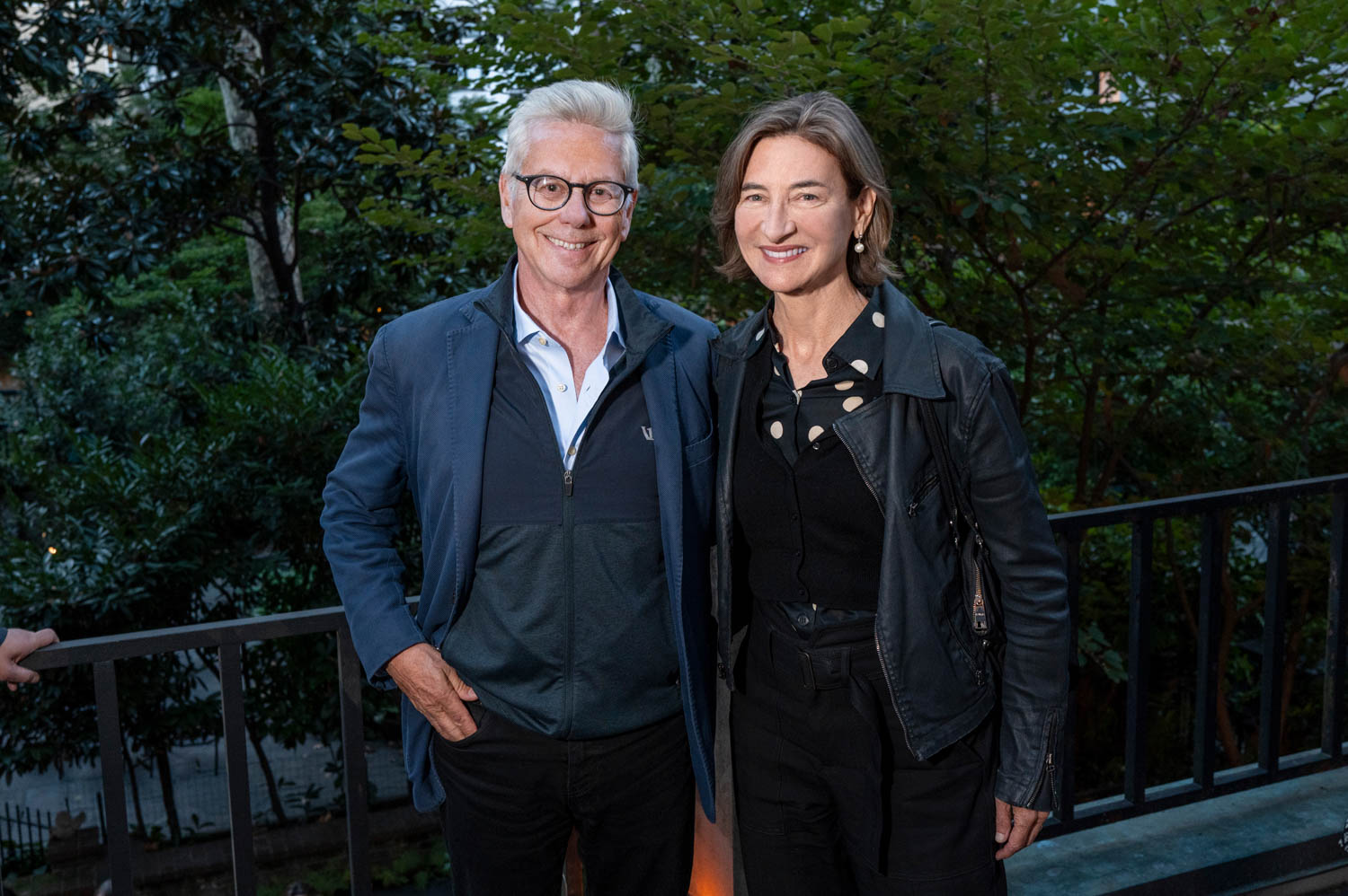 Two adults in jackets stand side by side and smile outdoors in front of a leafy background and metal railing.