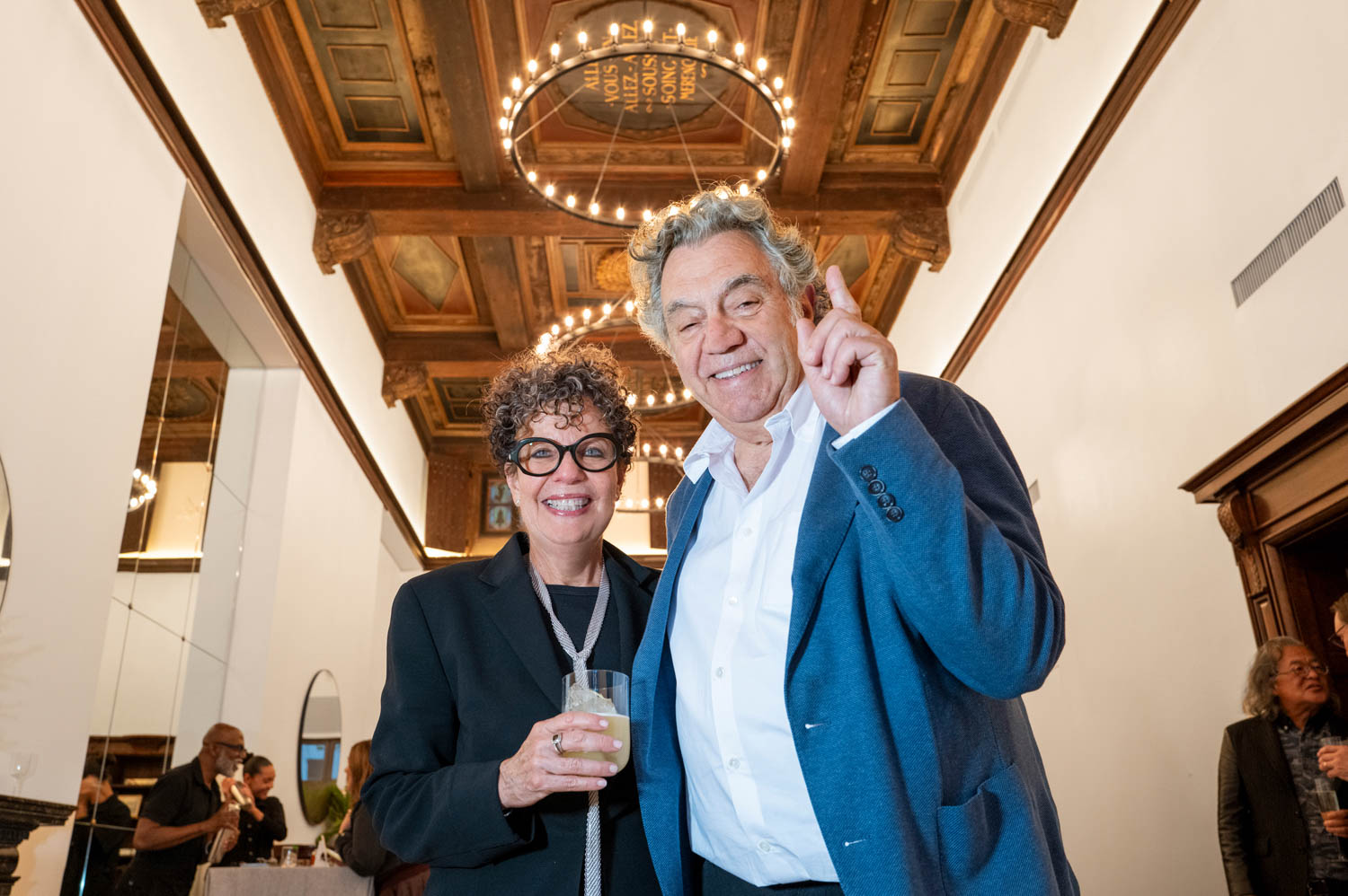 Two people dressed in business attire smile at the camera at an indoor event, with ornate ceilings and chandeliers visible above.