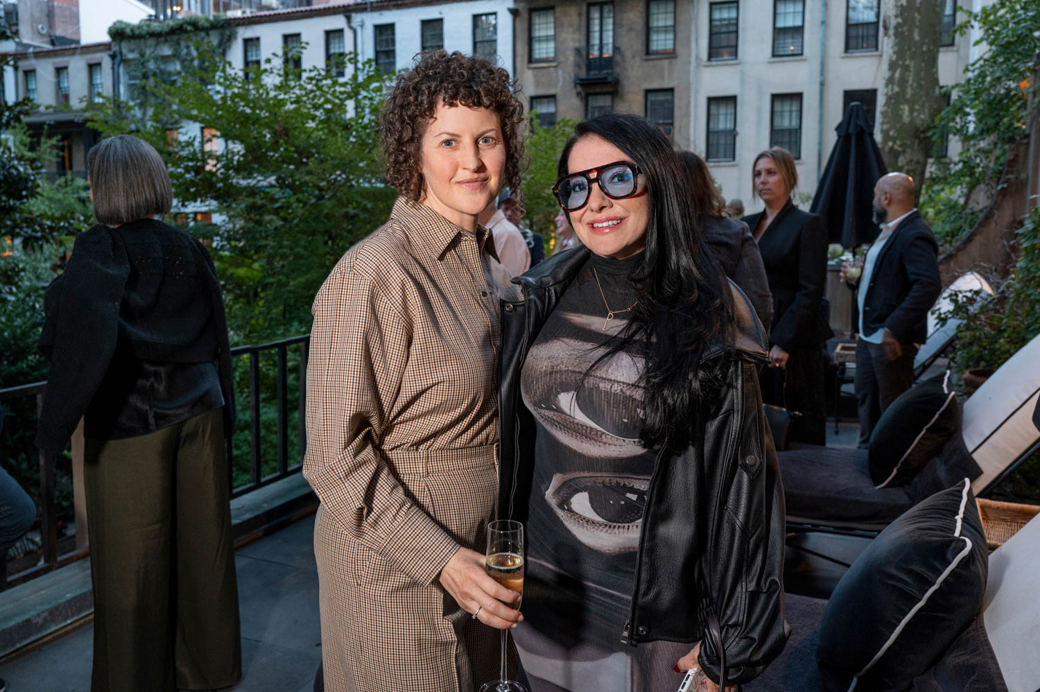 Two women stand together at an outdoor social event; one holds a drink, and people mingle in the background near urban buildings and greenery.