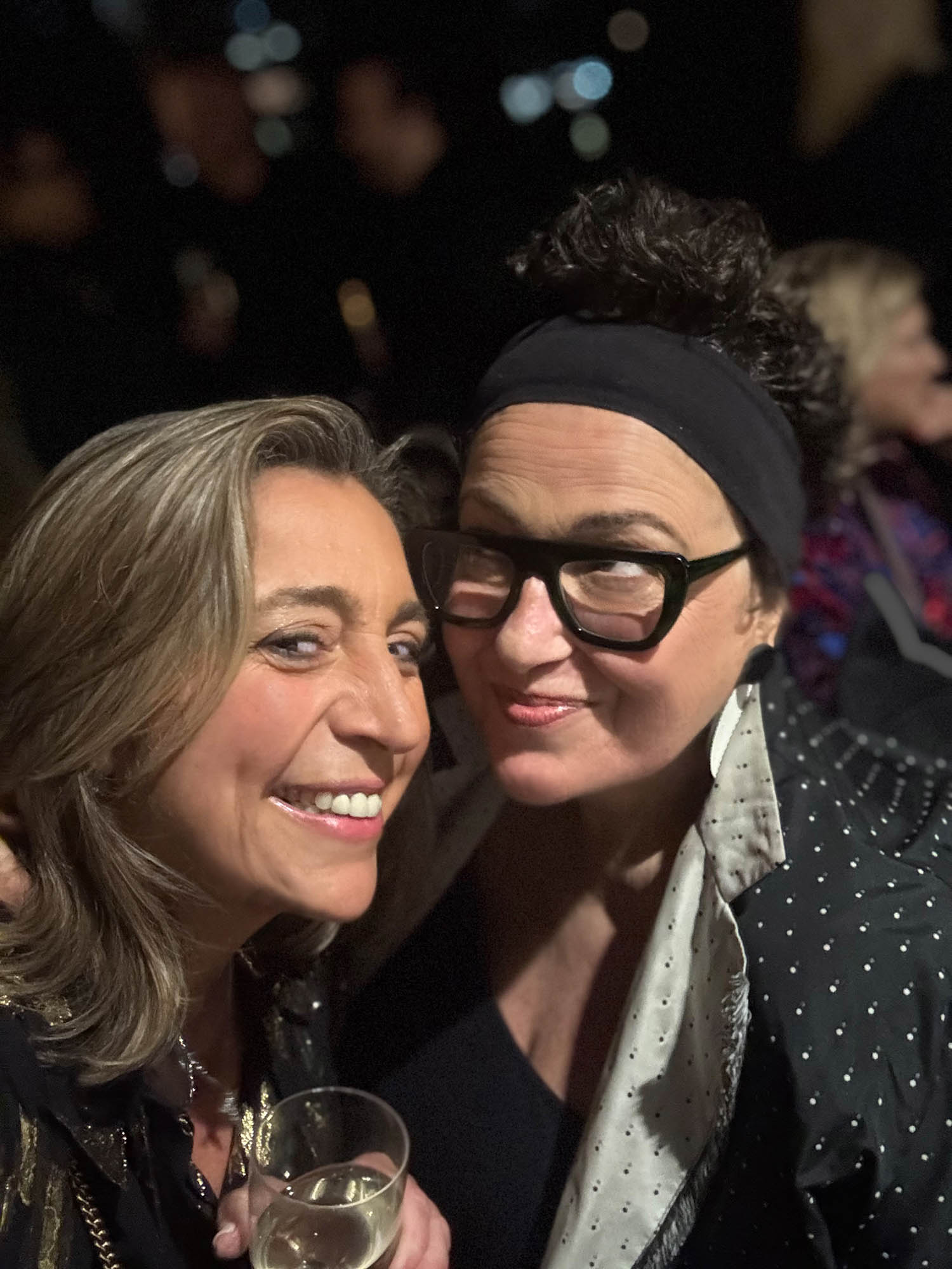 Two women pose closely together and smile at the camera during what appears to be a social event. One woman holds a glass of champagne. The background is dimly lit with other people visible.
