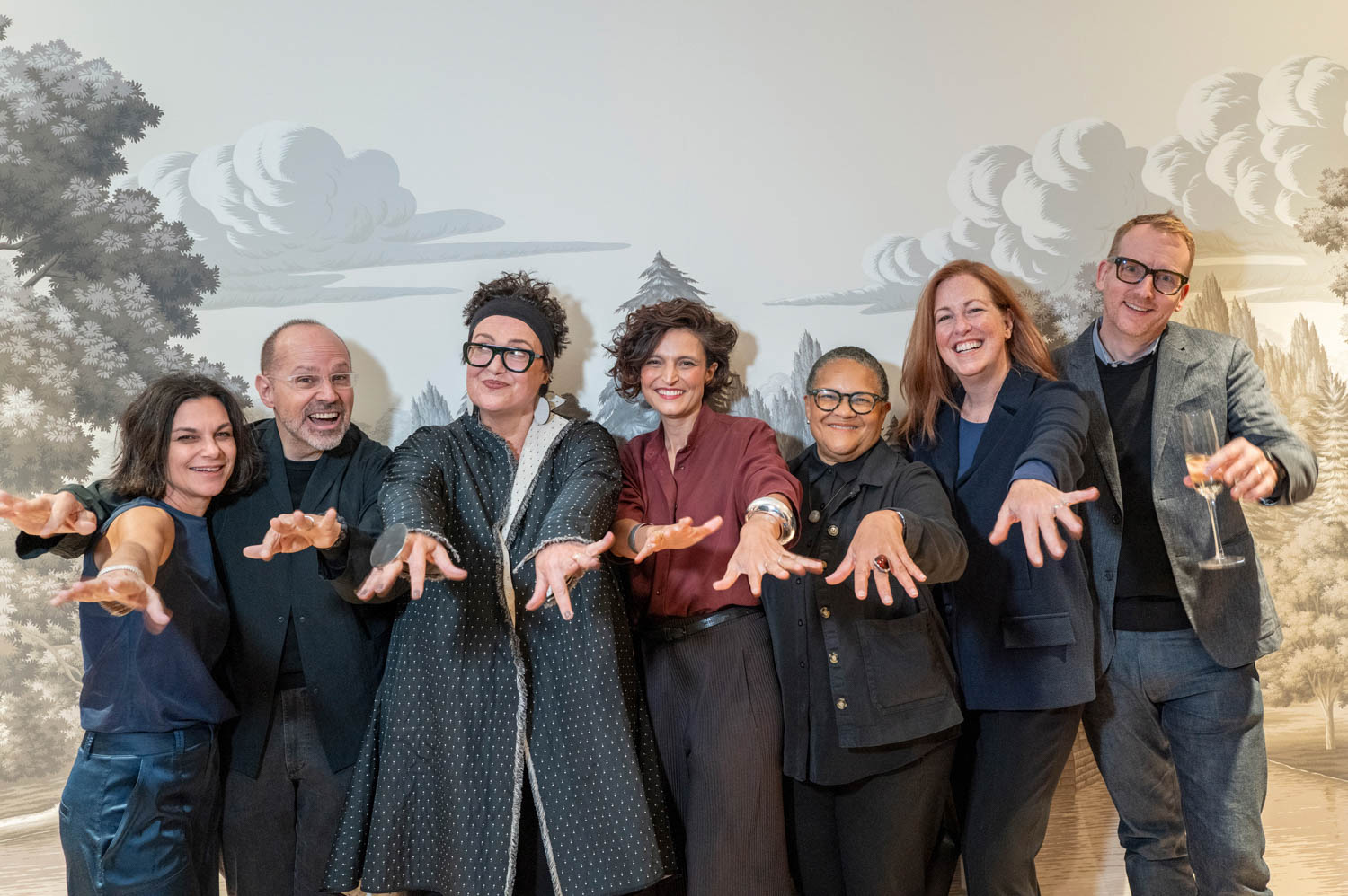 Seven people stand in a row indoors, smiling and displaying their hands toward the camera, with a painted landscape mural in the background.