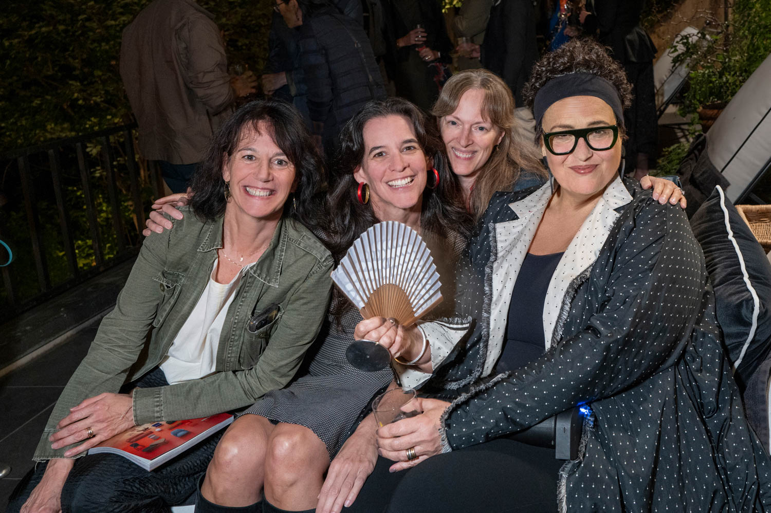 Four women sit closely together on a bench at an outdoor event, smiling at the camera. One woman holds a folded fan in her hand.