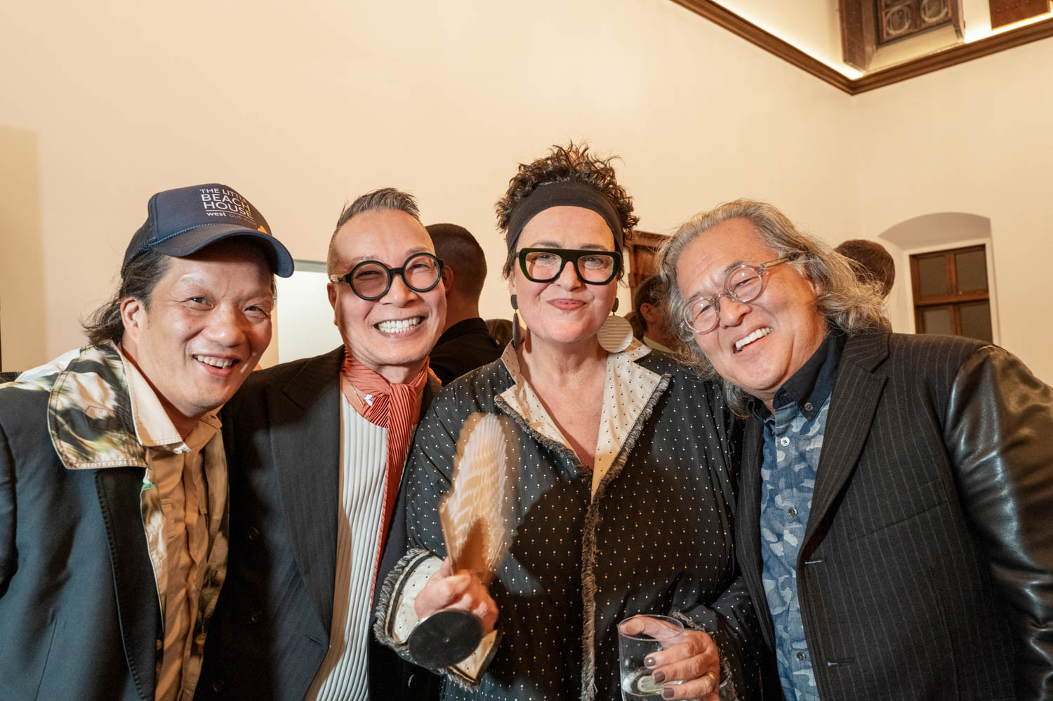 Four adults stand closely together indoors, smiling at the camera. One person holds a wooden paddle. They are dressed in stylish, eclectic outfits and glasses.