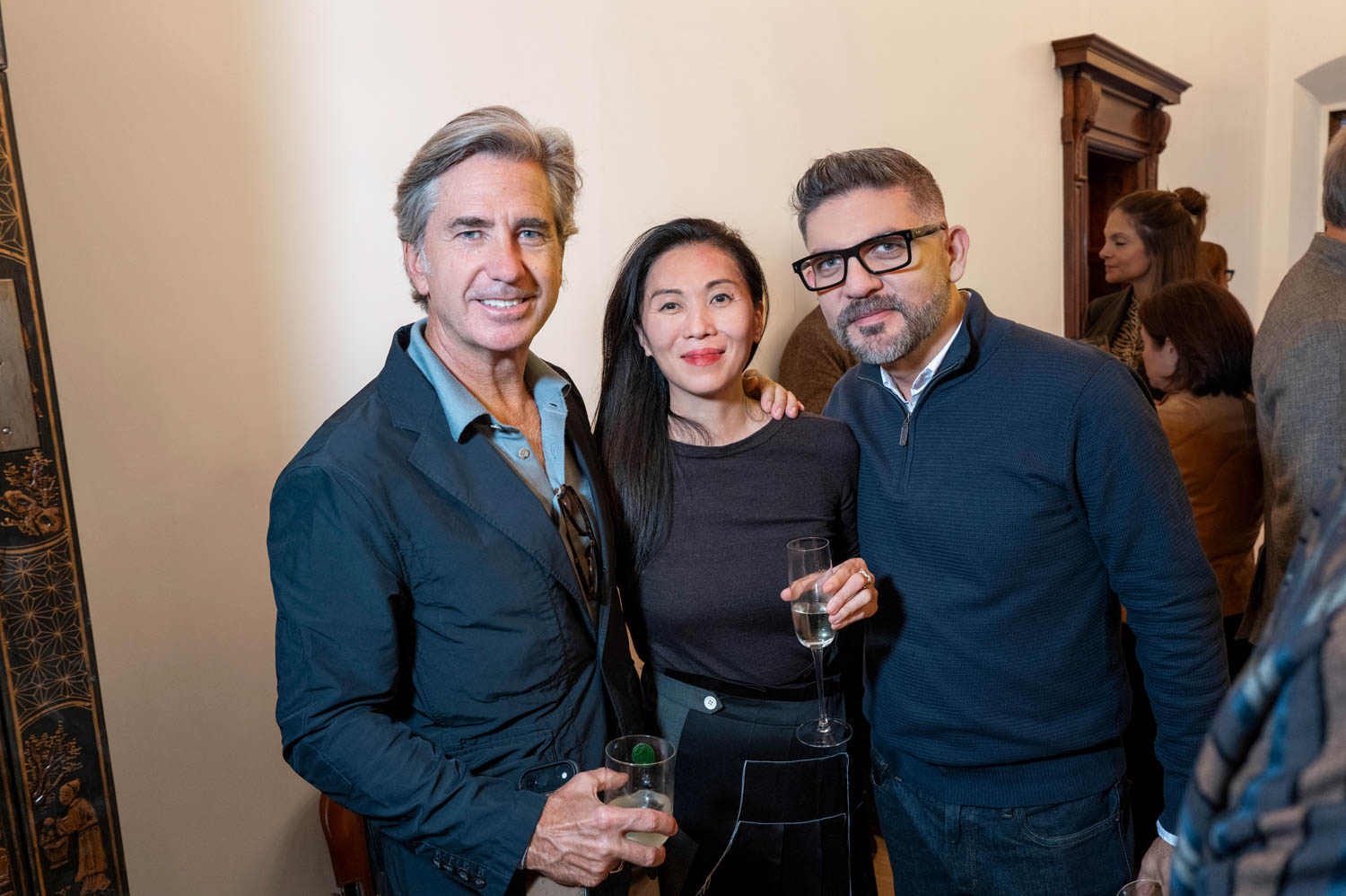 Three people pose indoors at a social gathering, holding drinks and smiling at the camera. Other guests are visible in the background.