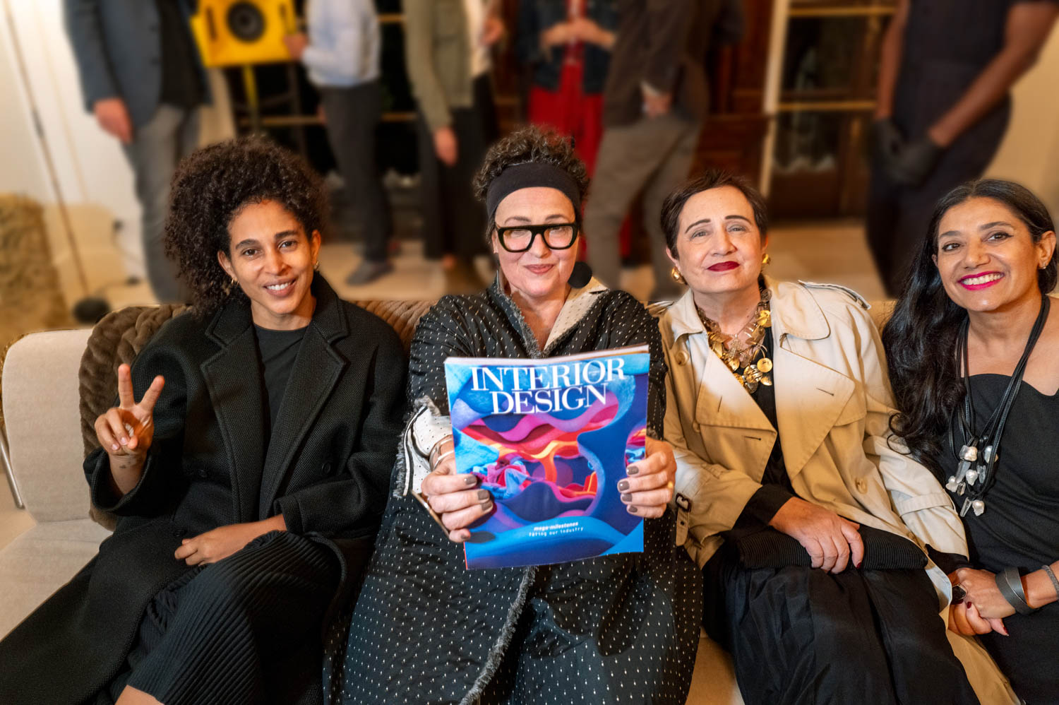 Four women sit on a couch; the woman second from left holds a magazine titled “Interior Design.” Several people stand blurred in the background.