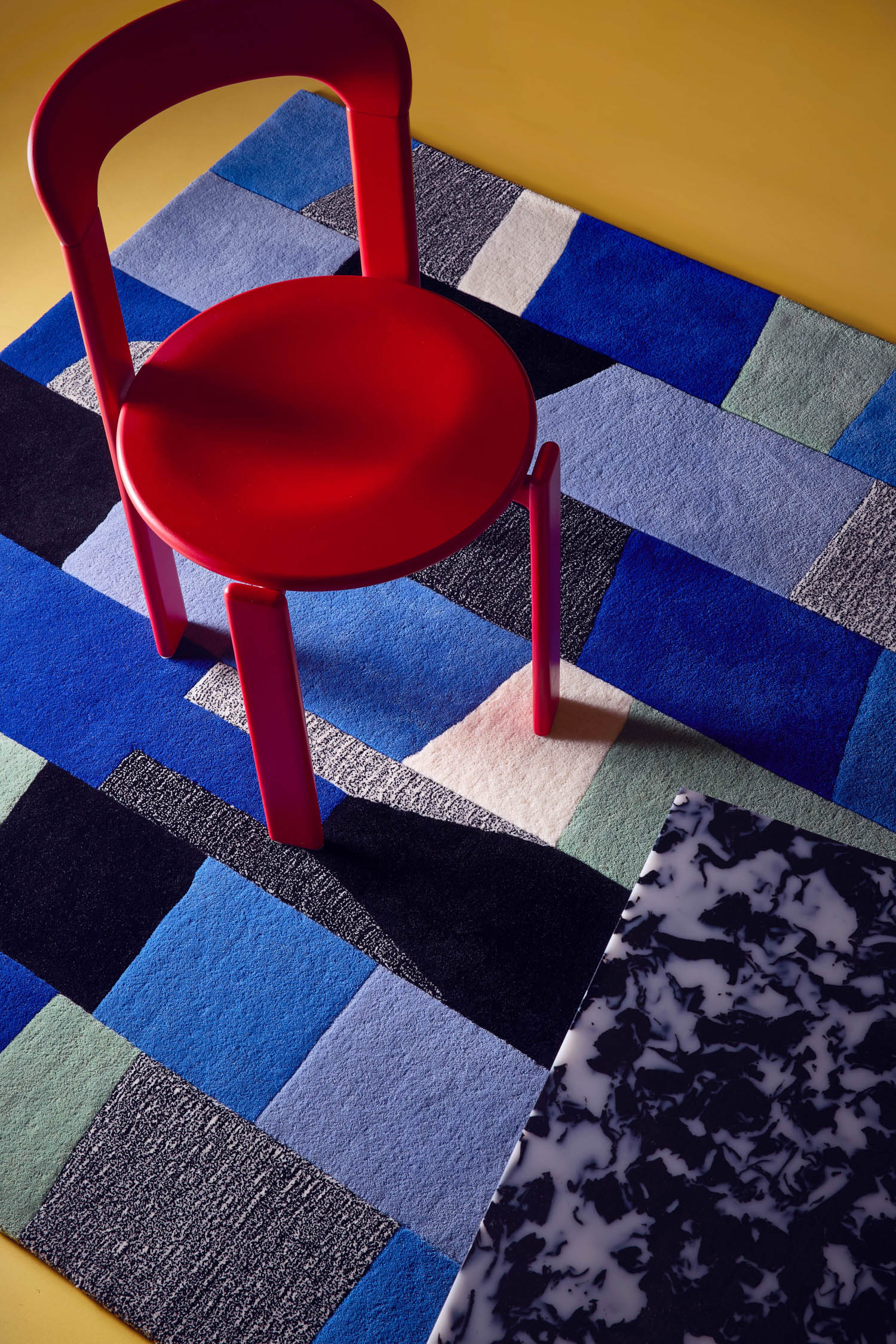 A red chair sits on a geometric blue, black, and gray rug next to a table with a black-and-white marbled surface, all on a yellow floor.