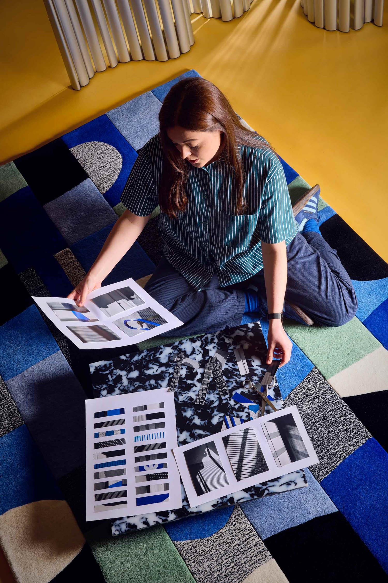 A woman sits cross-legged on a patterned rug, examining several sheets of printed geometric designs spread out in front of her.