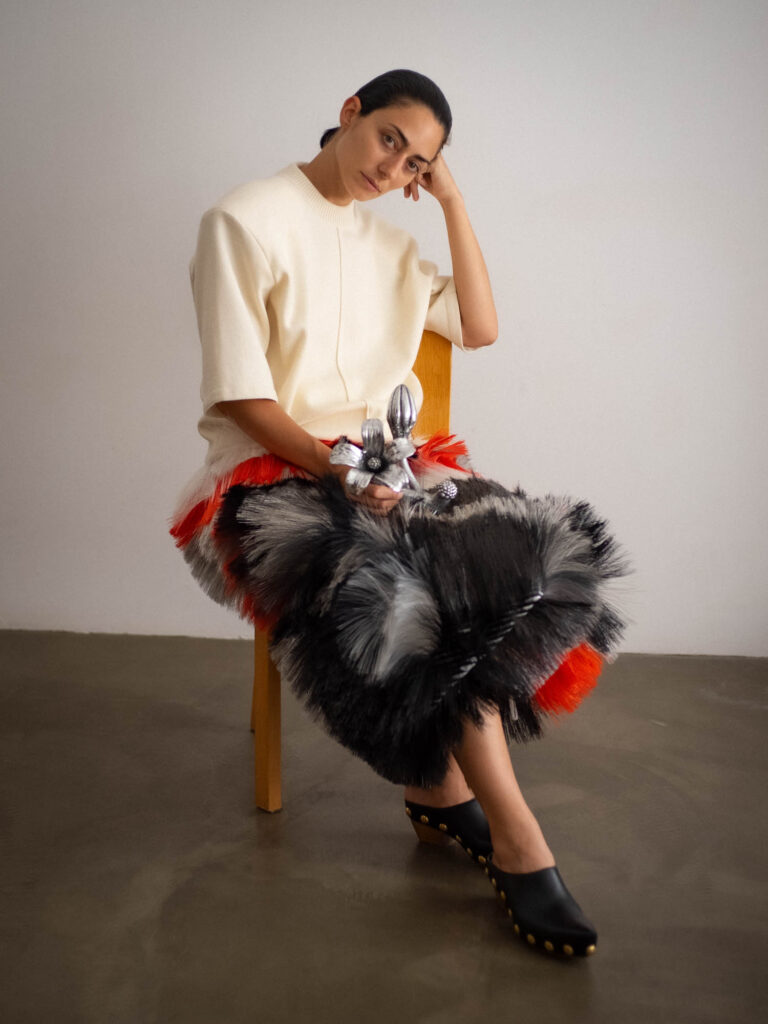 A person in a white top and a textured, multi-colored skirt sits on a wooden chair against a plain wall, resting their head on one hand.
