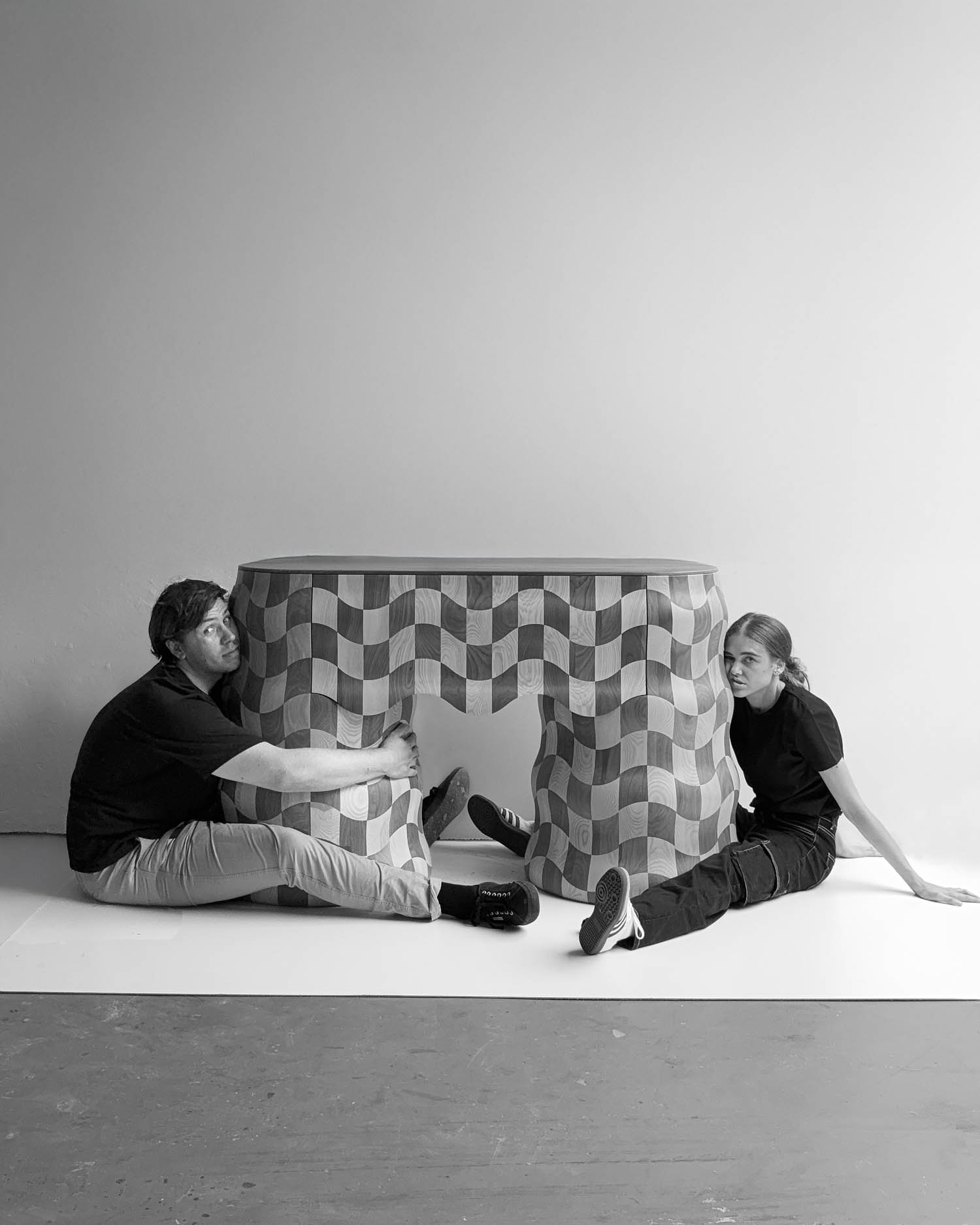 Two people in black shirts sit on the floor on either side of a geometric wooden table with a wavy, checkered pattern against a plain background.