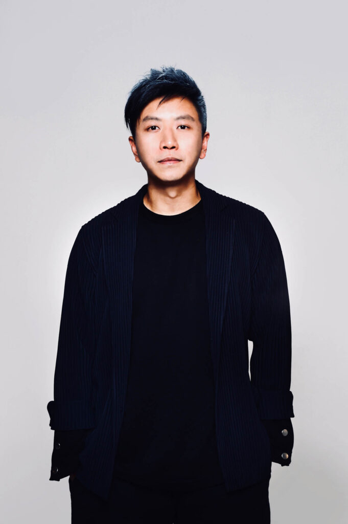 A person with short dark hair, wearing a black shirt and jacket, stands against a plain light background, facing the camera with a neutral expression.