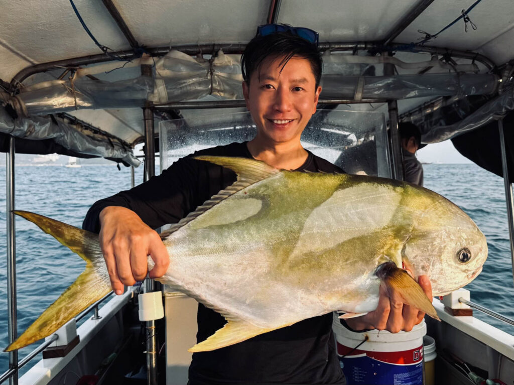Person on a boat holding a large fish with both hands, smiling at the camera; ocean and sky visible in the background.