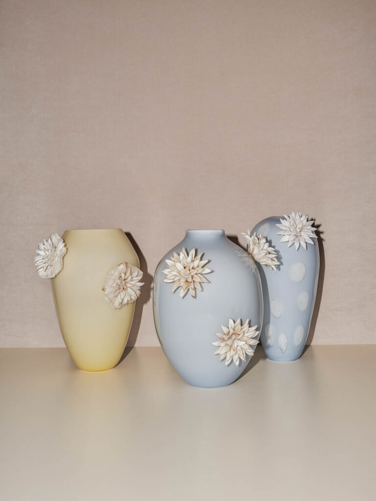 Three pastel-colored vases—yellow, blue, and blue with white dots—stand side by side, each decorated with white ceramic flowers, against a neutral beige background.