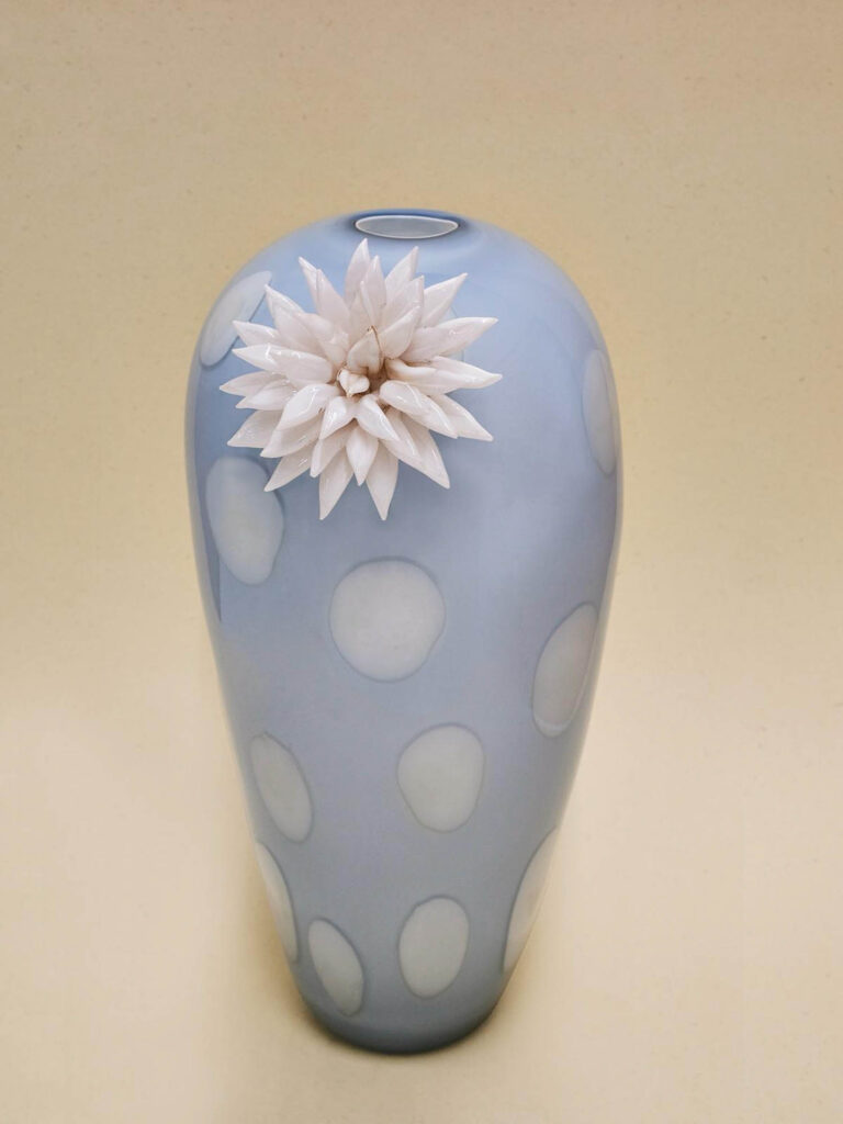Pale blue vase with large white polka dots and a white, spiky flower decoration near the top, set against a plain beige background.