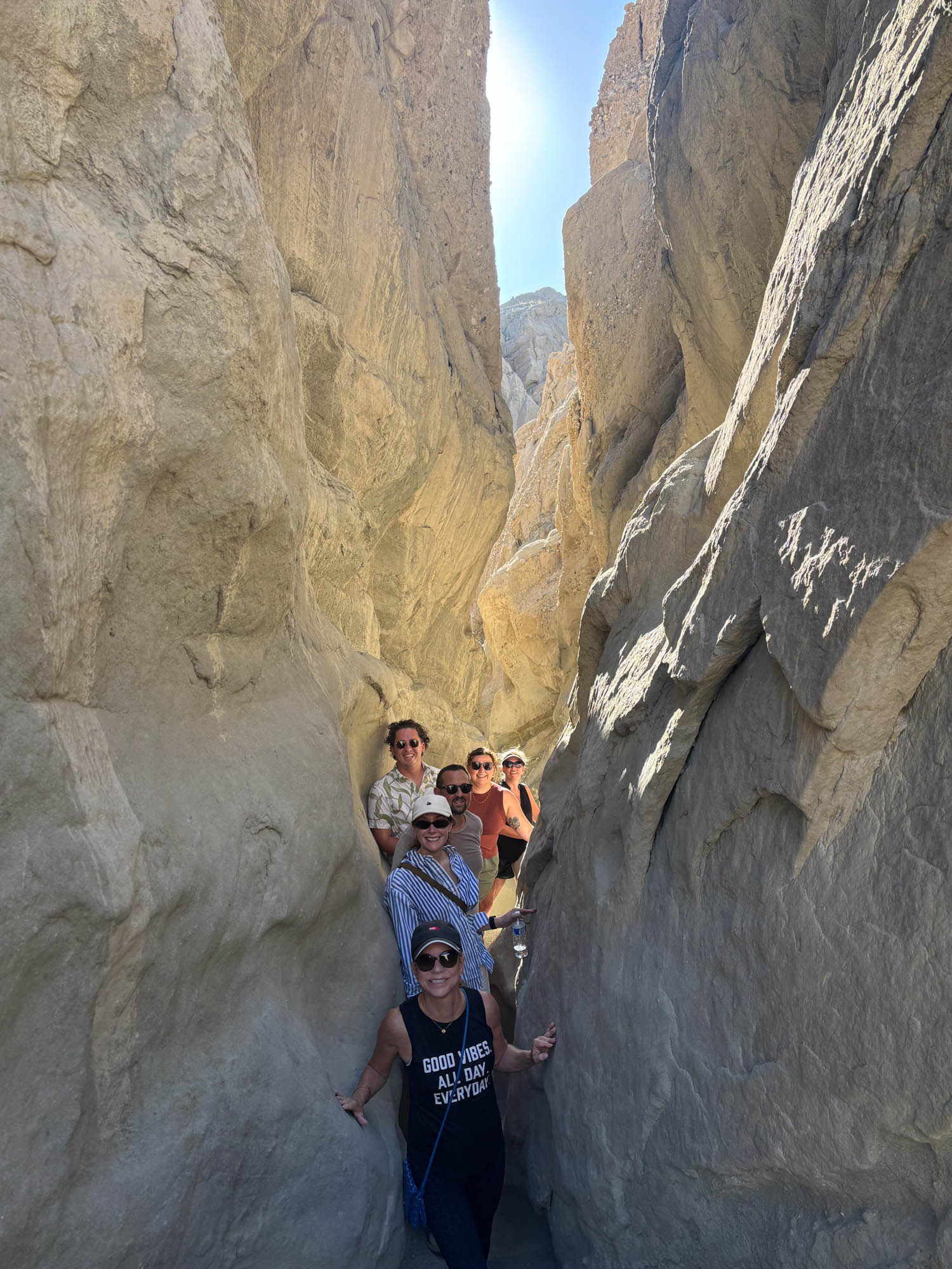 Six people standing in a narrow, sunlit rock canyon, posing for a group photo as they make their way through the passage.